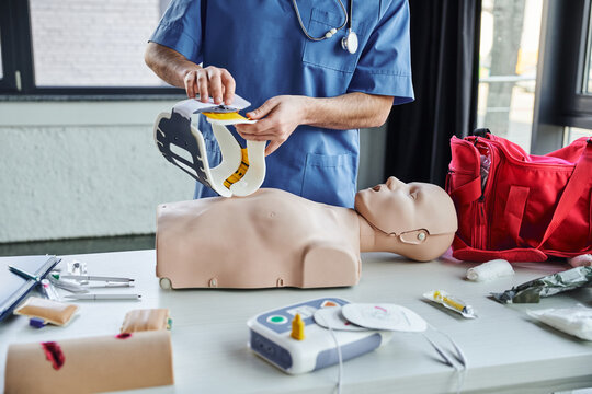partial view of healthcare worker in blue uniform holding neck brace above CPR manikin, first aid kit, defibrillator and medical devices in training room, life-saving skills development concept