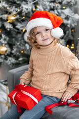 Portrait of a cute little girl in a beautiful knitted sweater ad a red Santa hat near decorated Christmas and new year tree at home. Smiling, cheerful and happy, holding red gift box