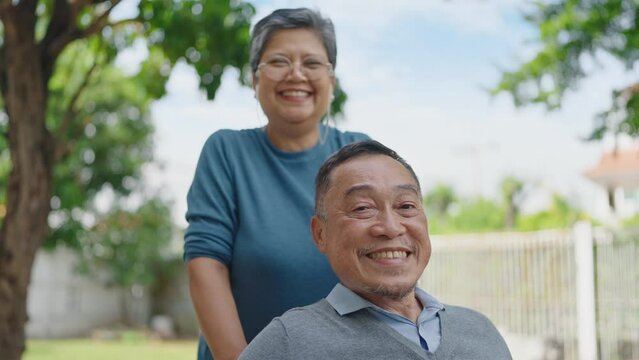 Cheerful asian adult male patient sitting on wheelchair and his wife smiling and looking at camera while relaxing together in the park. Relationship concept, Asian elderly married couple