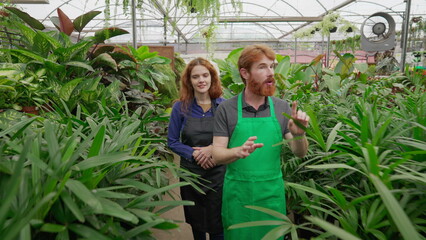 Obraz premium Two employees walking through isle inside plant store. man and woman staff wearing aprons, male employee explaining Flower Shop business