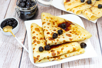 Homemade crepes with black wild bitter cherry jam, on a white ceramic plate, on a wood background, near a glass jar with. Home cooking