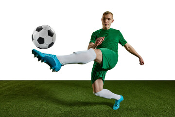 Competitive man, football player in green uniform training, kicking ball with leg against white...