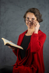 Mujer joven con lentes sonrriente leyendo un libro con un saco rojo y fondo gris