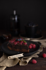 Still life with a piece of chocolate brownie cake with raspberries. Happy birthday inscription on the sign