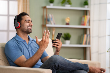 Cheerful indian man with wireless headphones listening favorite songs on mobile phone on sofa at...