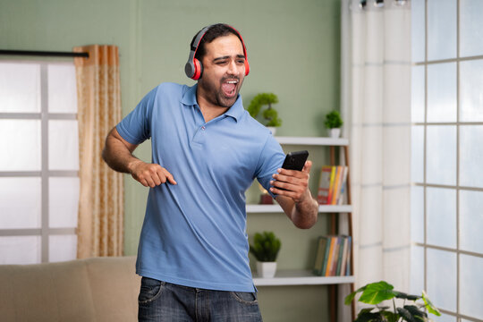 Cheerful happy indian man dancing while listening songs on headphones by holding mobile phone at home - concept of enjoyment, Weekend vibes and entertainment