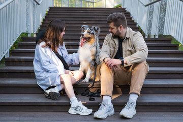 Happy young woman and man sitting with their aussie dog outdoors in urban park. Couple and their pet australian shepherd
