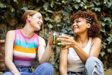 Two cute multiracial girls are happily toasting with some flavored cocktails in a place with vegetation. Concept of multiethnic couple drinking mojitos, toast with combined. - obrazy, fototapety, plakaty