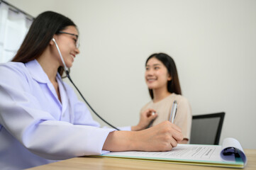 Focus of Asian female doctor hand checking heart rate of young female patient with stethoscope and taking note on prescription of doctors in the clinic