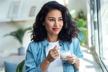 Happy beautiful woman eating yogurt while standing in living room at home.