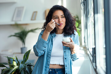 Happy beautiful woman eating yogurt while standing in living room at home.