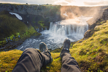 Legs of man sitting on edge of mountain with Axlafoss waterfall flowing in canyon at Icelandic Highlands