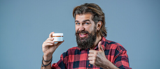 Bearded man with cream in his hands shows a thumbs up. Cream conditioner for men. Expert care for demanding hair. Plastic container with cream. Man holds bottle of cream or shower gel - obrazy, fototapety, plakaty