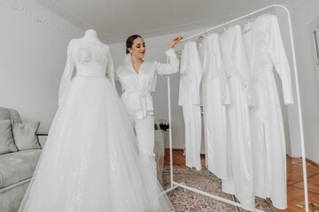 a beautiful bride poses in a white robe next to her wedding dress and her friends' dresses on a...