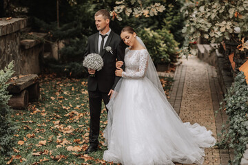Fototapeta premium A stylish couple of European newlyweds. A smiling bride in a white dress leaned against the groom's shoulder. Groom dressed in a classic black suit. Wedding in nature