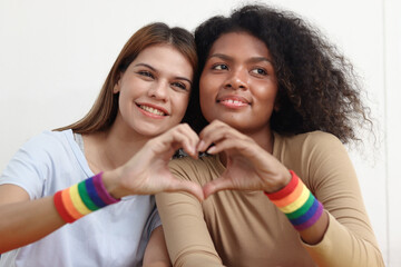 Happy smiling homosexual lesbian couple with rainbow flag wristband making heart sign by hand, beautiful woman and African curly hair girlfriend celebrate gay pride month together, romantic LGBT lover