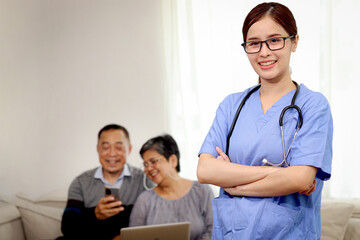 Portrait of happy smiling beautiful Asian woman doctor with stethoscope in blue coat standing with arms crossed in hospital with senior elderly patient couple as blurred background, medical worker.