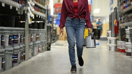 Woman shown from waist down, carrying can of paint and roller walking down paint aisle in hardware...