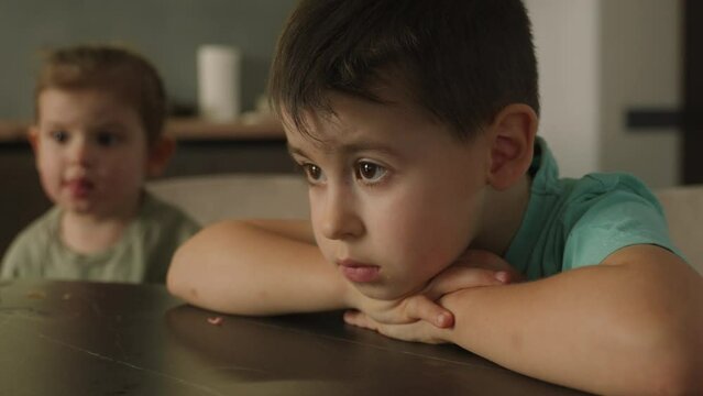 Cute little kid looking away while sitting in the kitchen at the table waiting food.