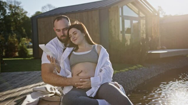 Happy man hugging pregnant woman near lake while watching ultrasound image
