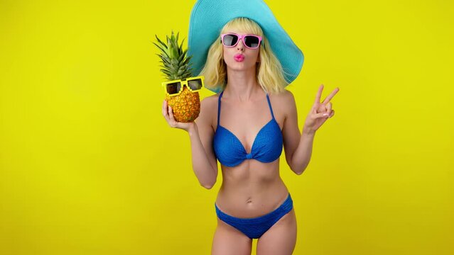 Sexy woman in bikini, peace hand, dancing with pineapple, fashionable hat, smiling enjoys summertime for spa, wellness. Playful slim lady dance for rest, tan or pool party, colorful trend on yellow
