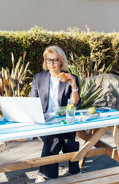 Middle-aged Businesswoman Having Lunch At The Park Outdoor Cafe, Picnic Area And Working On Laptop During Her Break. Balanced Healthy Diet Lunch Box. Unhealthy Eating Habits. Bad Work-life Balance.