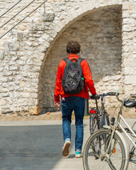 Fototapeta na wymiar A young man in a red jacket walks along an old European street with a bicycle and an electronic cigarette in his hand against the background of a stone medieval wall, healthy lifestyle and bad habits