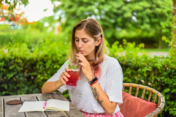 Cute  woman sitting on a chair in summer garden, drinking ice tea and writing in a notebook