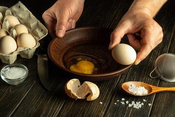 The cook is trying to break an egg on a plate before making an omelet. Egg breakfast by the hands of the chef on the kitchen table