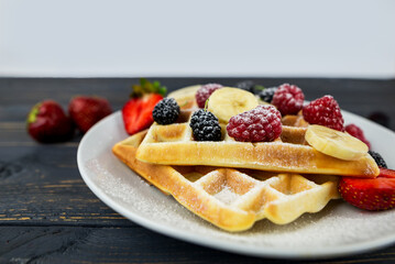 Belgian waffles with summer berries and powdered sugar in a white plate on a dark wooden background.