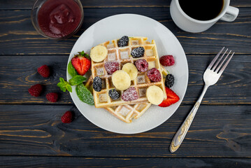 Belgian waffles with summer berries and powdered sugar in a white plate on a dark wooden background.