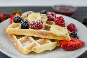 Belgian waffles with summer berries and powdered sugar in a white plate on a dark wooden background.
