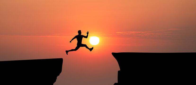 Concept of reaching life and business goals. Silhouette of man jumping over chasm at sunrise
