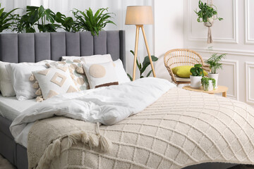 Comfortable bed, wicker armchair, lamp and beautiful houseplants in room. Bedroom interior
