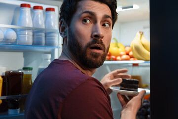 Man taking plate with cake from refrigerator in kitchen at night. Bad habit