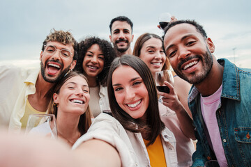 Big group of young adult happy friends smiling taking a selfie portrait and looking at camera with friendly expression. A lot of cheerful multiracial people celebrating and laughing. Buddies bonding