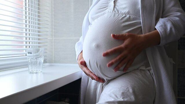 Close-up of pregnant woman, gravid female, expectant mother in white nightgown, stroking her big belly in late pregnancy time, standing by window in a cozy home interior. People. Maternity. Lifestyle