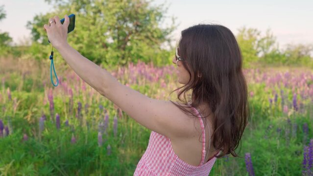 Young beautiful traveler woman take selfie on phone in the pink flower garden. Smiling attractive girl in white sunglasses filming herself on cellphone in lupine flowers field. Relaxing on outdoor