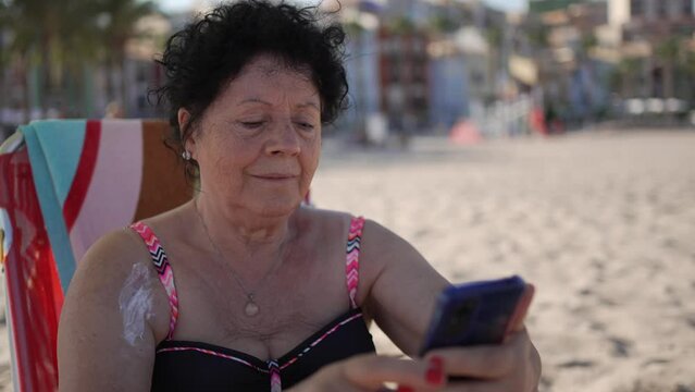 Old woman on the beach with her mobile phone. Video of an elderly woman under the umbrella manipulating her smartphone.