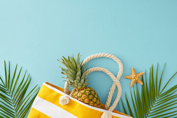 Concept of a beachside tropical vacation. Top view shot of striped beach bag, ripe pineapple, palm leaves and starfish on pastel blue background with empty space for promo or text