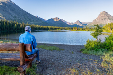 Man, person sitting watching a scene of a lake surrounded by trees and a rugged mountain in the...
