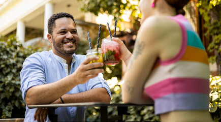 A multiethnic couple toast with mojitos happily while sitting in a place with vegetation. The woman is unrecognizable, celebrating success with cocktails with alcohol.