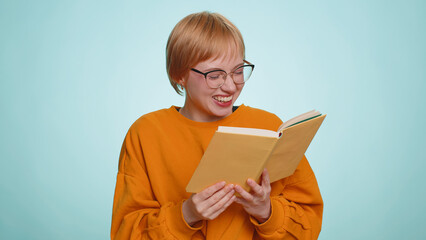 Young woman in glasses reading funny interesting fairytale story book, laughing, leisure hobby, knowledge wisdom, education, learning, study, wow. Blonde pretty girl isolated on blue studio background