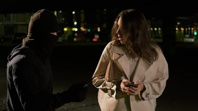 A woman stands at a bus stop with a handbag uses the phone. A masked robber attacks with a knife and takes away a smartphone and handbag. Robbery