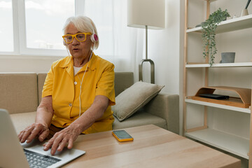Happy elderly woman with a laptop typing in headphones smile sitting at home on the couch in a...