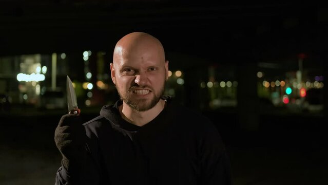 A bald Caucasian young criminal in black clothes looks menacingly at the camera with a grin of his teeth. Holds a gloved knife in his hands and opens it in an attempt to attack