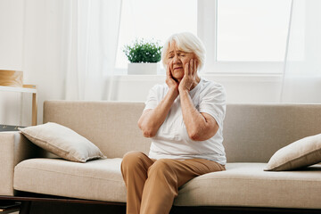 Elderly woman severe pain in the head sitting on the couch, health problems in old age, poor...