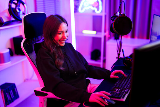 Asian pretty gamer girl typing keyboard and use mouse while sitting ergonomic chair and playing video game challenge in entertainment neon light room