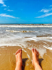 Bare feet on the beach sand, lapping the sea water with white foam and blue sky.