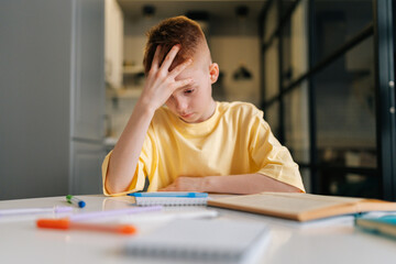 Low-angle view portrait of exhausted pupil boy tired from studying holding head head with hand sitting at desk with paper notebook, sad looking down. Frustrated child schoolboy doing homework at home.
